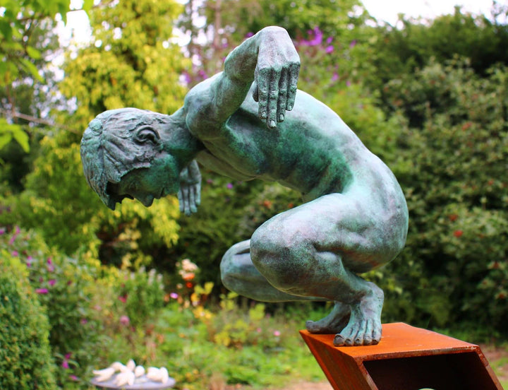 Large bronze sculpture of a crouching male figure, arms extended and head bowed, balanced on the edge of an angled rusted steel plinth. The figure’s green patinated surface contrasts with the warm, textured tones of the steel base. Muscular detail and tension in the pose evoke focus, strength, and introspection. Displayed both indoors against gallery walls and outdoors in a garden setting, the work captures light and shadow across the form, emphasising anatomical realism and expressive movement.
