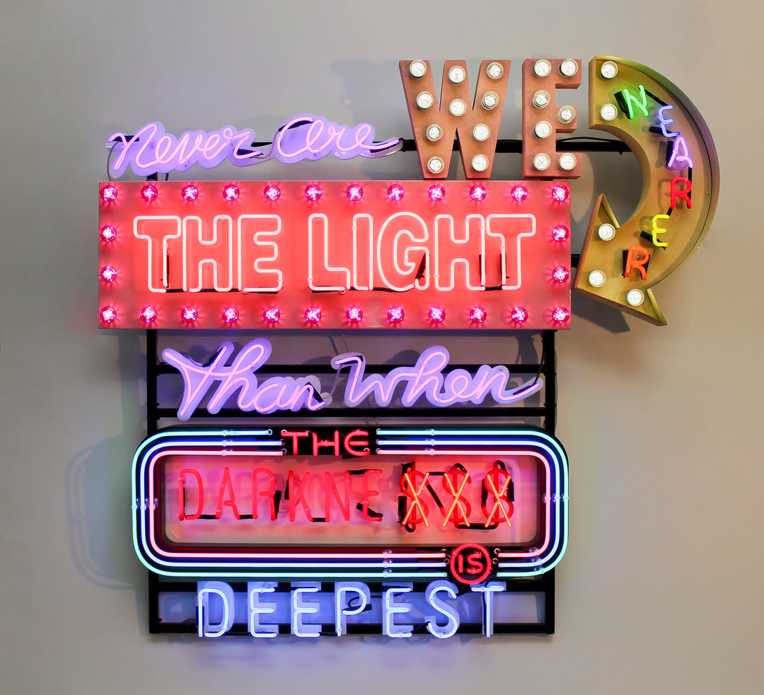 Neon wall artwork featuring the phrase “Never Are We Nearer The Light Than When The Darkness Is Deepest” in layered typography. Includes fabricated metal panels, red marquee lights, a retro arrow lit with cabochon LEDs, and vivid neon lettering in magenta, violet, red, blue, turquoise, orange, and white. A glowing, multicoloured composition by Chris Bracey reflecting spiritual insight and emotional depth.