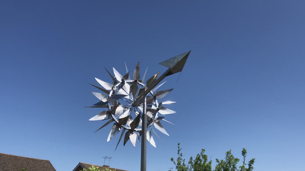 Intricate kinetic sculpture by Martin Jarvis composed of multiple polished stainless steel blades arranged in a radial formation, mounted on a vertical pole with a directional arrow. The piece rotates smoothly with the wind, casting shifting reflections and creating a mesmerising visual effect.