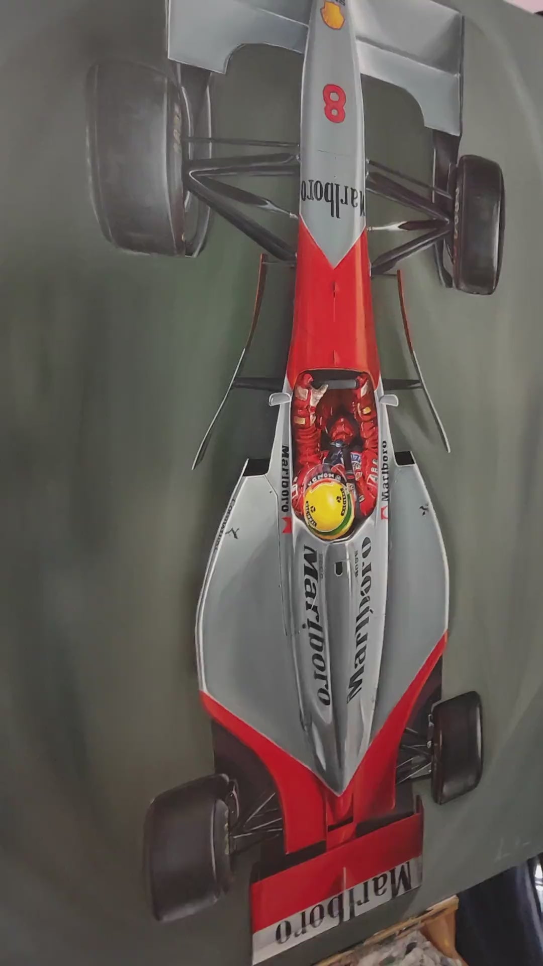 Video of Hyper-realistic top-down painting of Ayrton Senna driving the legendary McLaren MP4/4 Formula 1 car, finished in iconic Marlboro red and white livery, set against a deep grey racing track background. Senna’s bright yellow helmet glows at the centre of the sleek aerodynamic body, capturing speed, control, and motorsport heritage in a striking aerial perspective.