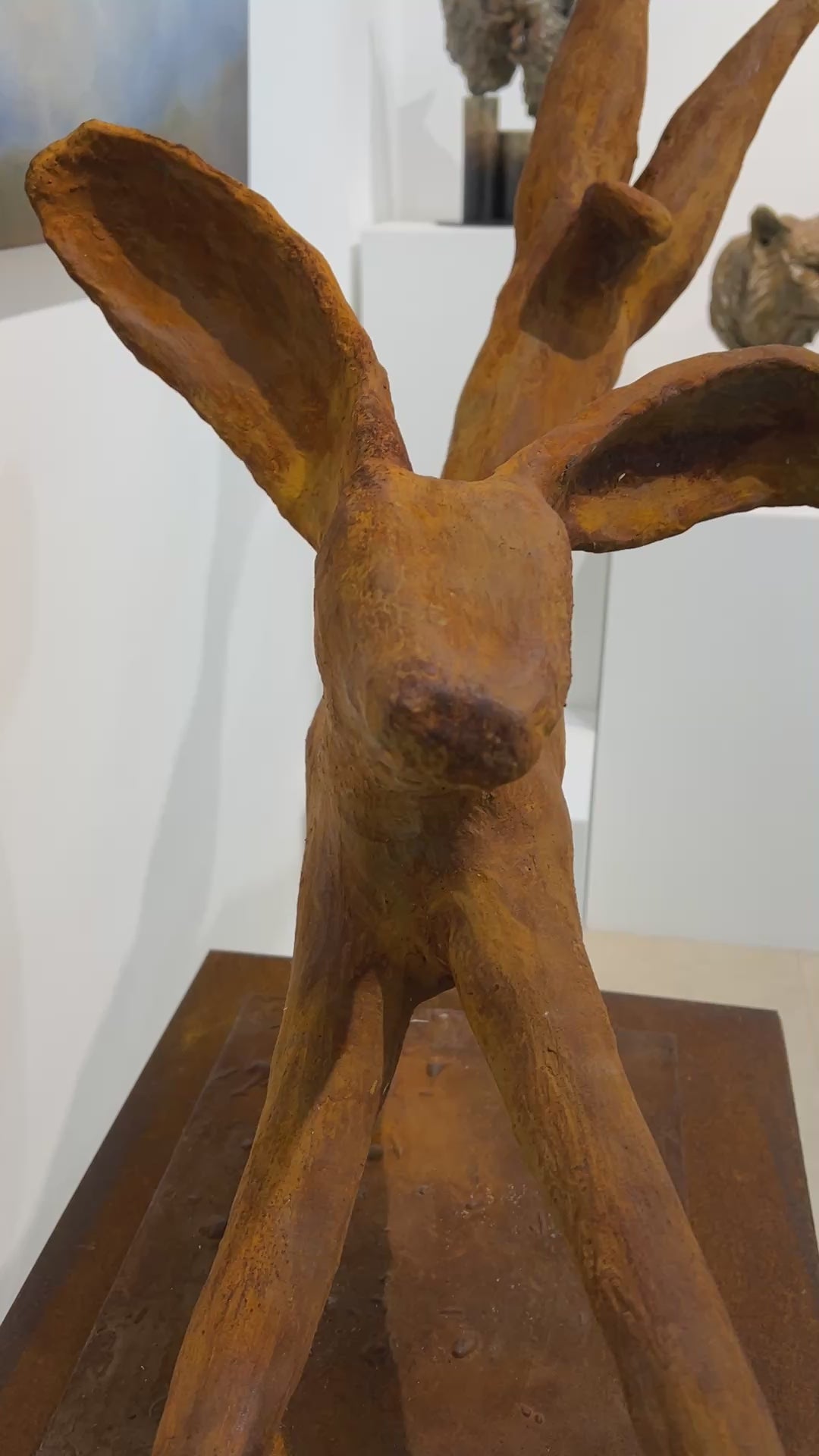 Freedom Majestic Hare sculpture by Min Reid, dynamic leaping hare captured mid-flight in warm bronze toned iron resin, mounted on a wooden base with countryside landscape, green fields and blue sky with white clouds in the background