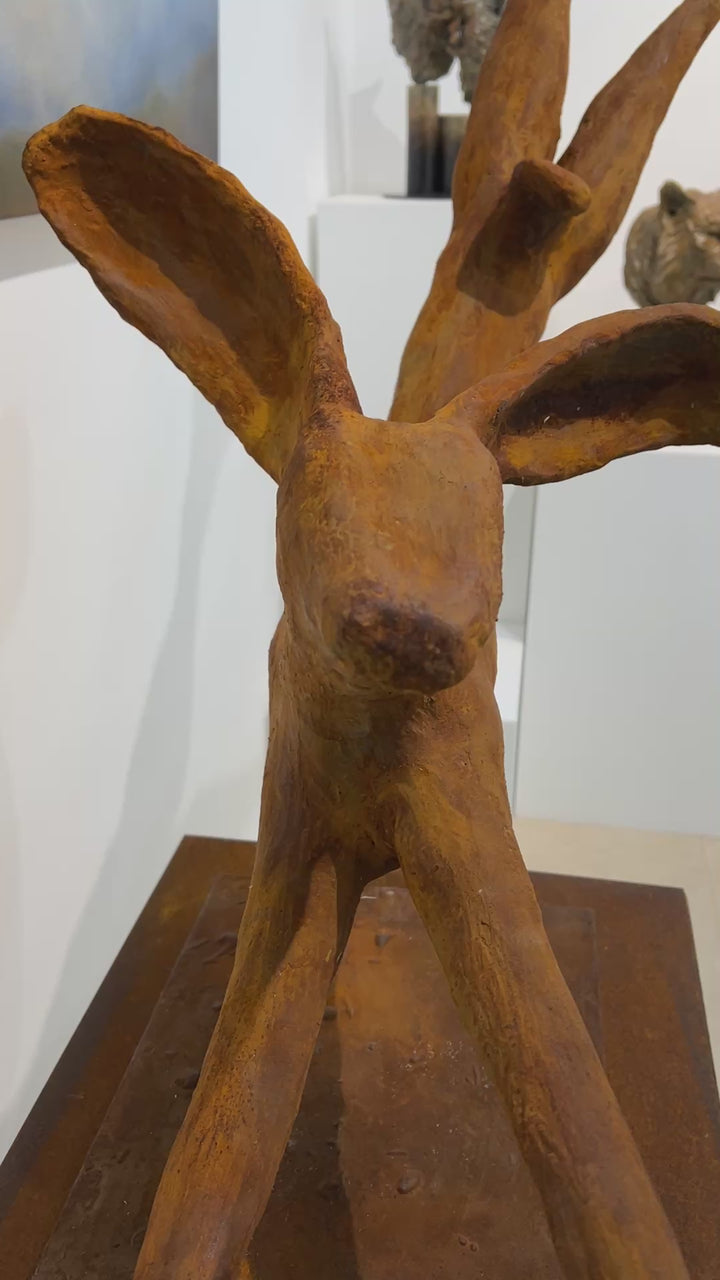 Freedom Majestic Hare sculpture by Min Reid, dynamic leaping hare captured mid-flight in warm bronze toned iron resin, mounted on a wooden base with countryside landscape, green fields and blue sky with white clouds in the background
