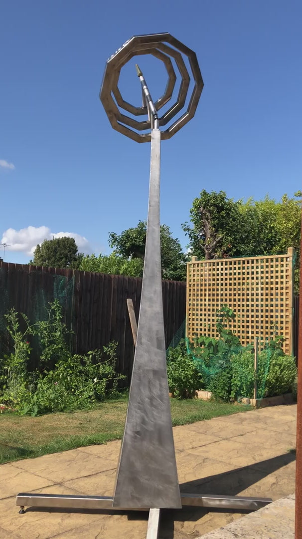 Martin Jarvis kinetic sculpture titled Serenity, featuring interlocking octagonal rings crafted from polished stainless steel. The rings are mounted atop a tall, tapered plinth and rotate gently in the breeze, creating a mesmerising visual rhythm that contrasts geometric precision with organic movement.