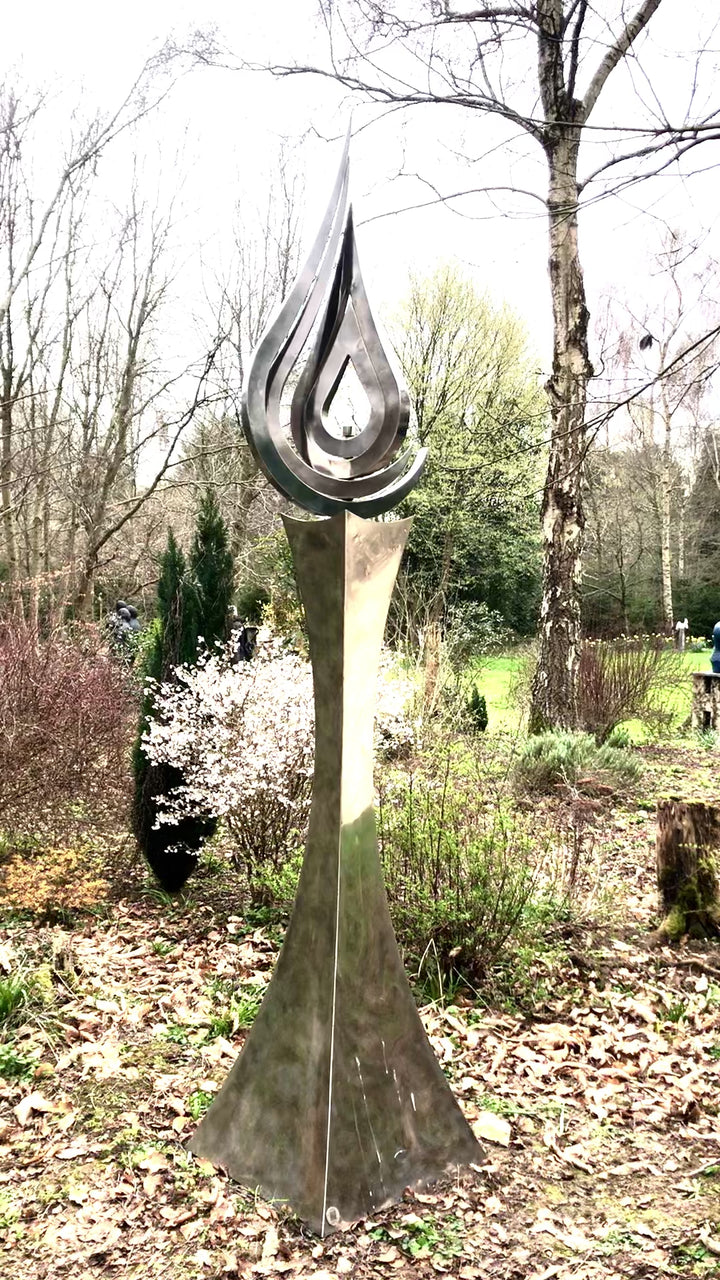 An elegant stainless steel kinetic sculpture by Martin Jarvis, designed in the form of a tall flame-like spire. The sculpture’s upper structure features fluid, ribbon-like curves that twist upward and move gently with the wind. Polished to a mirror finish, the steel reflects light and surroundings, giving it a dynamic presence. The base is solid and tapering, with soft surface patterning that adds depth and texture. Installed outdoors in natural and garden settings, the piece feels both modern 