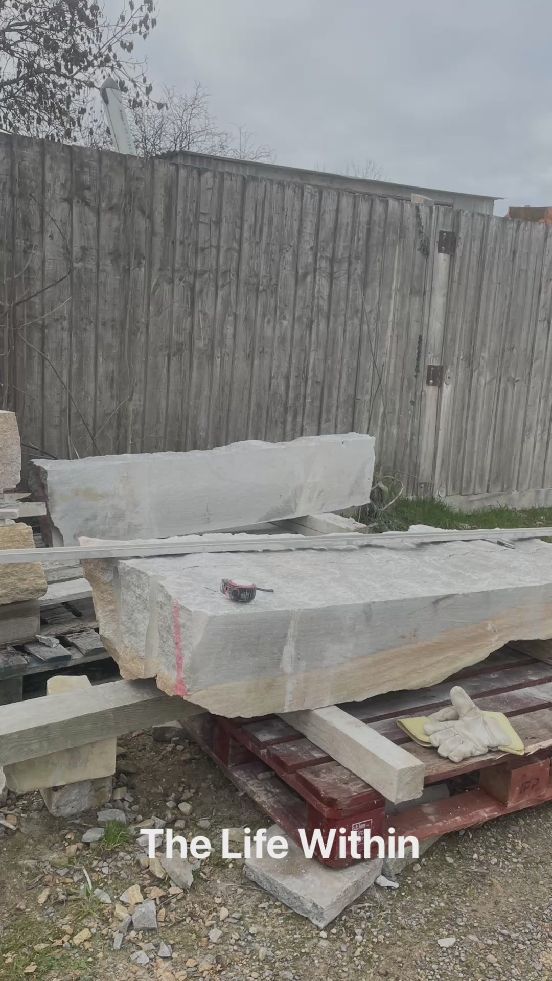 Tall grey Ancaster stone sculpture by Martin Cash with carved hollow centre and flowing white vein motif, mounted on a natural stone base in a landscaped garden setting.