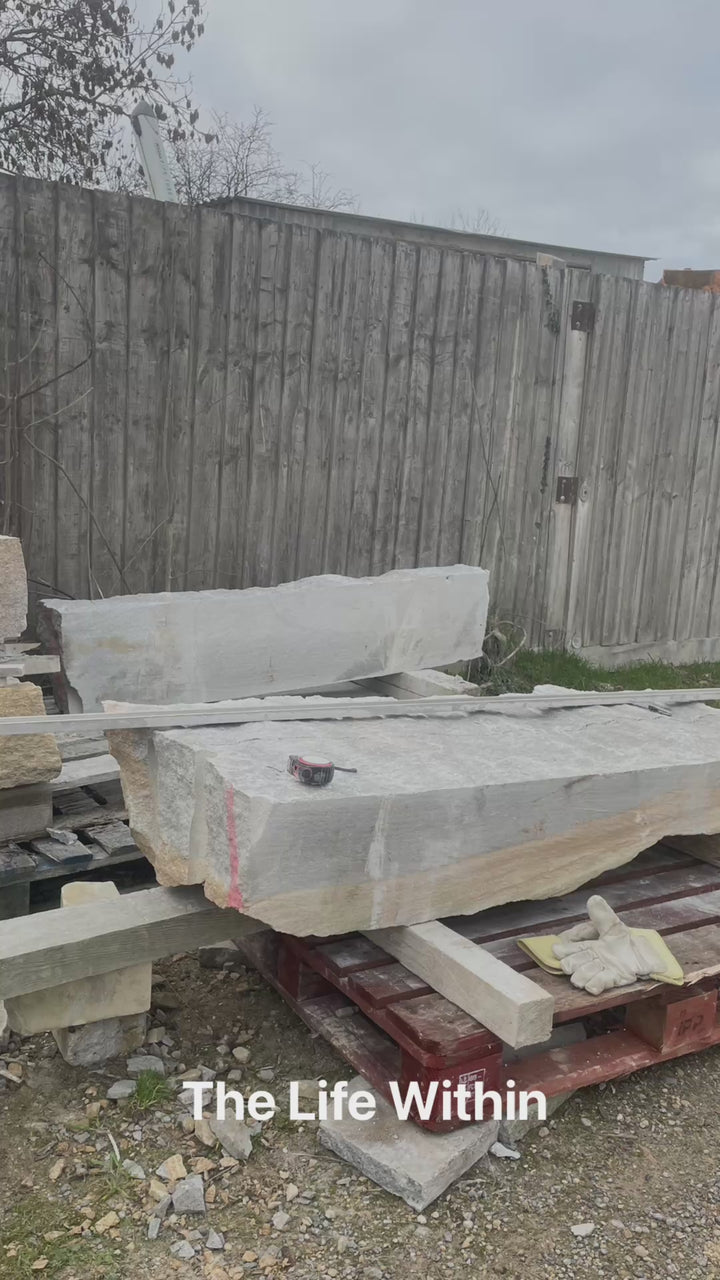 Tall grey Ancaster stone sculpture by Martin Cash with carved hollow centre and flowing white vein motif, mounted on a natural stone base in a landscaped garden setting.