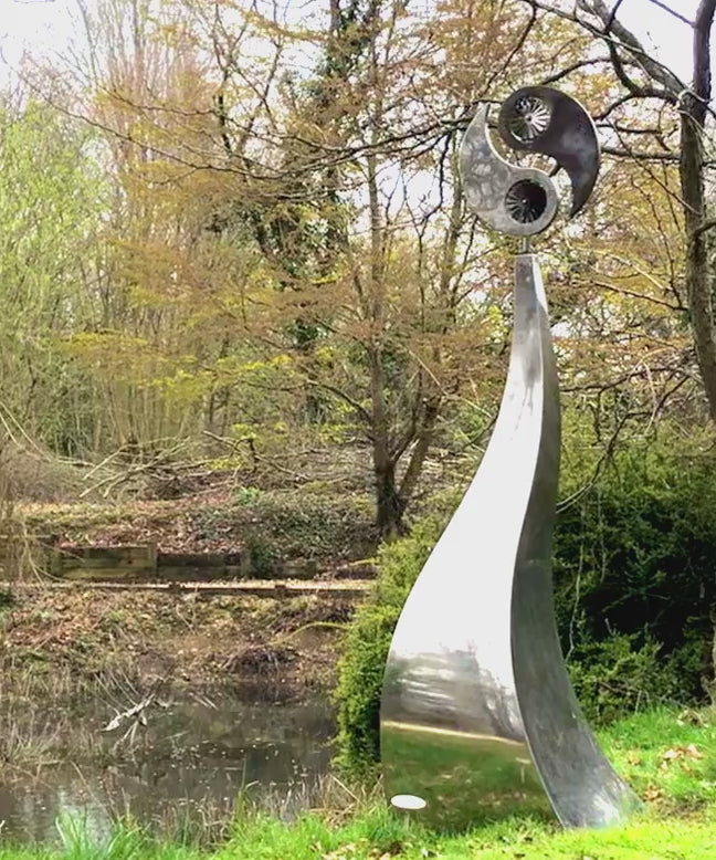 Polished stainless steel kinetic sculpture by Martin Jarvis featuring a tall, elegantly curved base topped with a dual-disc structure in motion. The upper element evokes a stylised yin-yang form, incorporating integrated fans that spin gently with the wind, creating a continuous, meditative rhythm.