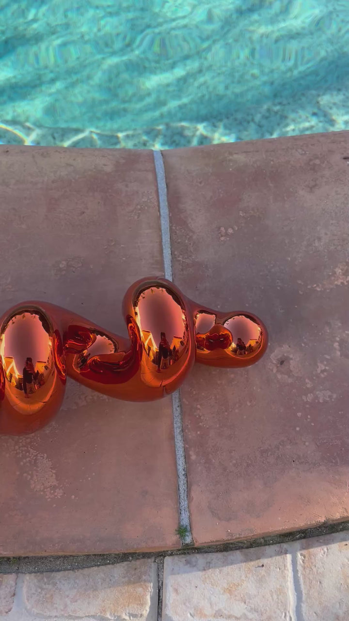Freestanding sculptural artwork spelling ‘LOVE’ in a flowing, cursive form, finished in high-gloss orange-tinted stainless steel.