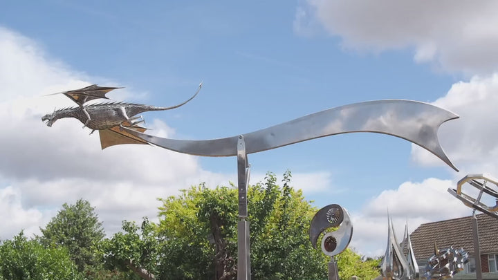 An impressive kinetic dragon sculpture by Martin Jarvis, made entirely from polished and textured metal. The dragon is mounted on a sweeping, curved arm that allows it to pivot and move with the wind. The body is detailed with overlapping metal scales, while the expansive wings—crafted from dark, matte-finished panels—give the illusion of flight. The sculpture captures dynamic energy and motion, designed to gently sway or turn with air currents. Positioned outdoors against blue skies, the piece