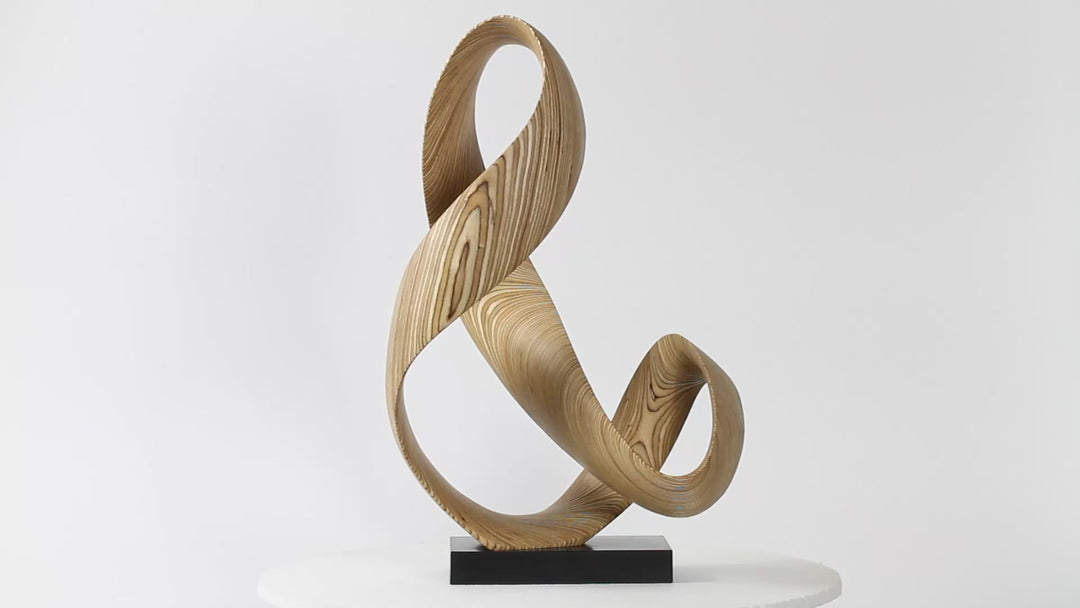 Hand-carved birch plywood sculpture by Bill Prickett, formed as a flowing Möbius-style infinity loop inspired by an ampersand, with continuous twisting curves, visible layered grain, and a balanced sculptural presence on a white base.