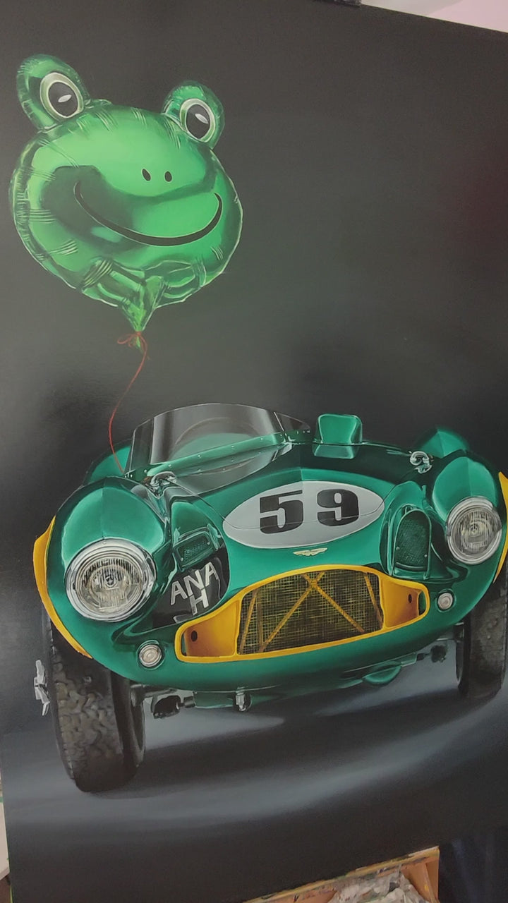 Video of hyper-realistic painting of a classic British racing green Aston Martin DB3S front-on against a deep black background, with a shiny metallic green frog-shaped balloon floating above the car, its red string tied to the vehicle — a playful contrast of automotive heritage and childlike whimsy.