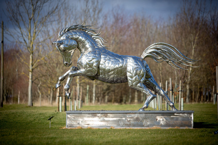 ‘Leaping Horse’ by Michael Turner, a unique, one-of-one stainless steel equestrian sculpture depicting a magnificent life-size horse leaping forward with its front legs raised and hind legs extended. Handcrafted from marine-grade stainless steel, the sculpture features intricately welded metallic plates, a powerful muscular body, flowing mane and sweeping tail. Its polished silver finish reflects the surrounding landscape, while the dynamic pose captures the strength, elegance and movement of a horse in ful
