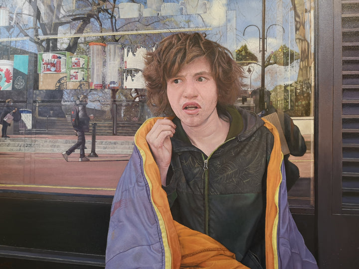 Rory by James Earley — a hyperrealist oil painting portraying a homeless teenage boy seated outside a London storefront, wrapped in a weathered coat with expressive eyes and an uncertain expression, while the window behind him reflects passing pedestrians and commercial branding, symbolising a world moving on without him.