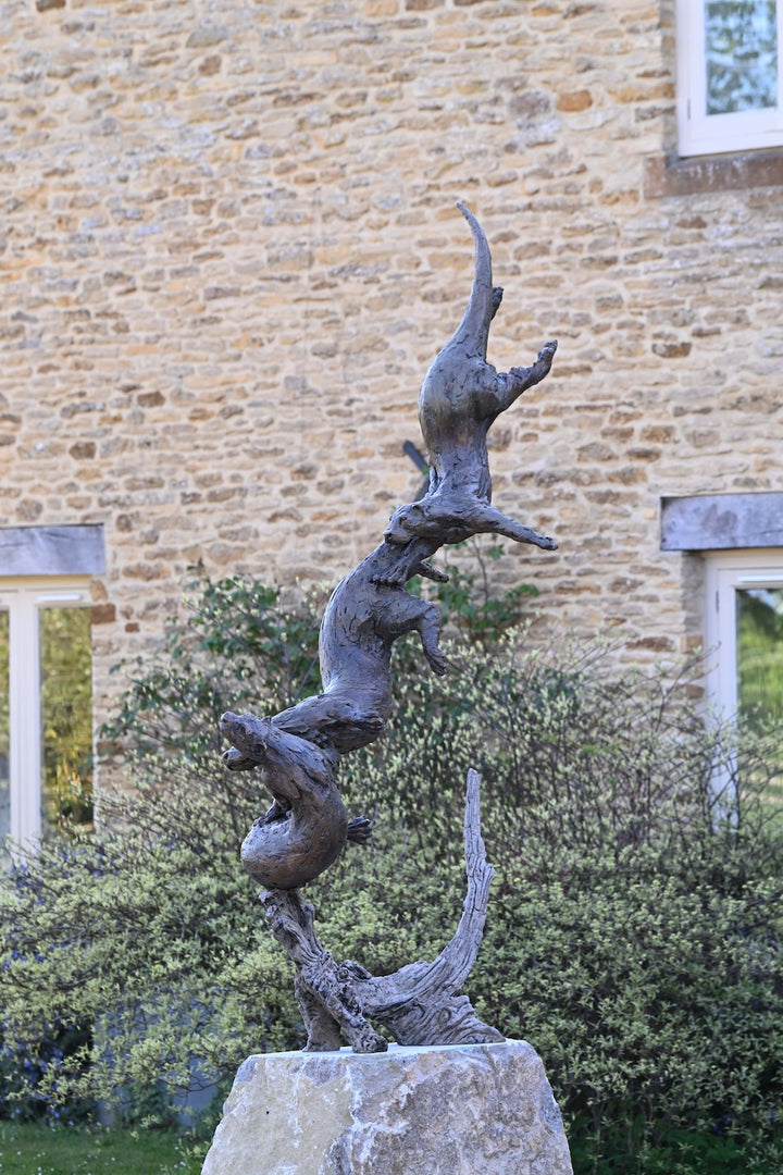 ‘Otters Playing Underwater’ by Hamish Mackie, a life-size limited-edition bronze wildlife sculpture depicting a mother otter and her playful cubs twisting, diving and swimming around a sculpted underwater tree root. The dynamic vertical composition features expressive animal movements, detailed fur textures and a weathered bronze finish in rich brown and earthy tones, capturing the playful energy and natural behaviour of otters beneath the water.