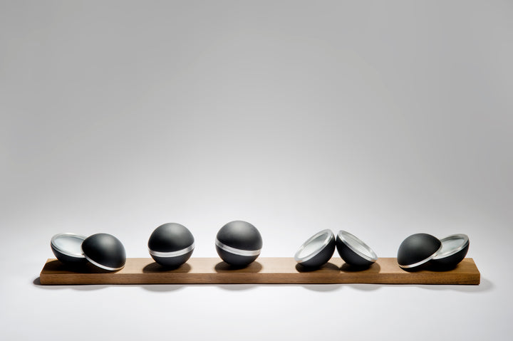 Serendipity by Tracy Nicholls — a unique glass sculpture of five black lead crystal spheres resting on a walnut base. Each sphere is formed from four lost wax cast hemispheres, the opaque black outer shells contrasting with luminous interiors of layered coloured glass. Aluminium rings and concealed neodymium magnets connect the hemispheres, allowing them to open and reveal swirling patterns of colour created by chance during the casting process. The polished surfaces and precise engineering contrast with th