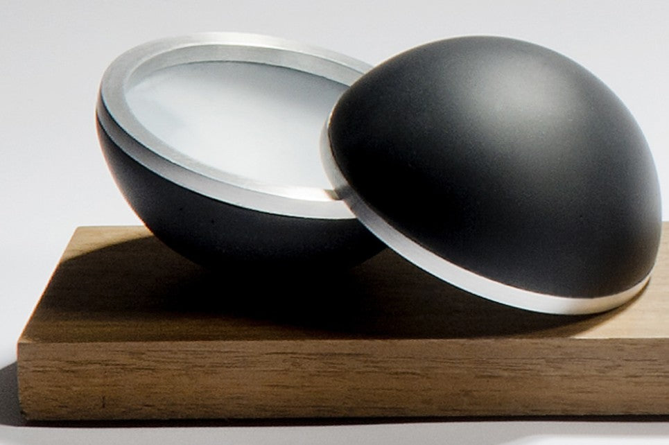 Serendipity by Tracy Nicholls — a unique glass sculpture of five black lead crystal spheres resting on a walnut base. Each sphere is formed from four lost wax cast hemispheres, the opaque black outer shells contrasting with luminous interiors of layered coloured glass. Aluminium rings and concealed neodymium magnets connect the hemispheres, allowing them to open and reveal swirling patterns of colour created by chance during the casting process. The polished surfaces and precise engineering contrast with th