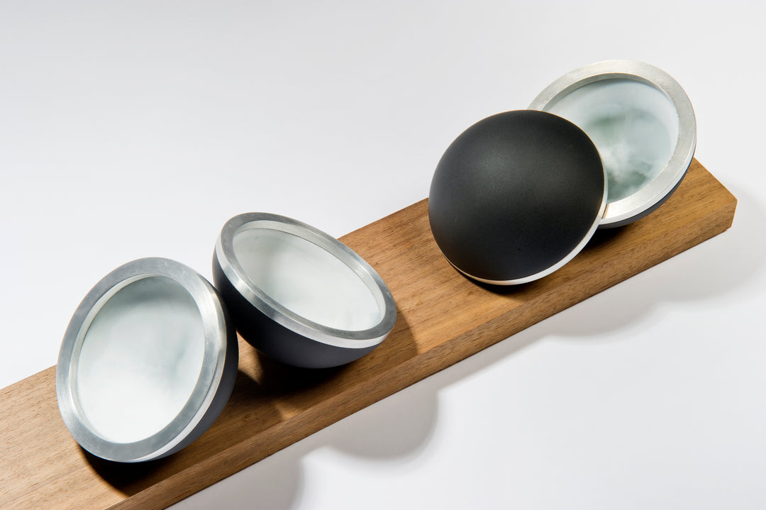 Serendipity by Tracy Nicholls — a unique glass sculpture of five black lead crystal spheres resting on a walnut base. Each sphere is formed from four lost wax cast hemispheres, the opaque black outer shells contrasting with luminous interiors of layered coloured glass. Aluminium rings and concealed neodymium magnets connect the hemispheres, allowing them to open and reveal swirling patterns of colour created by chance during the casting process. The polished surfaces and precise engineering contrast with th