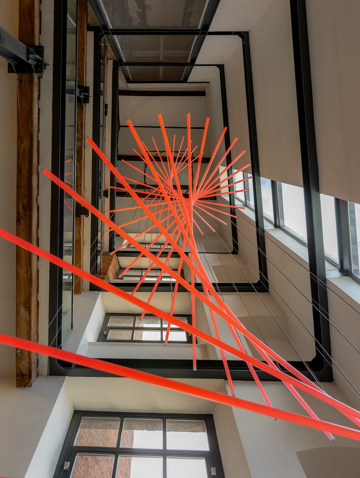 Suspended spiral light installation composed of intersecting illuminated linear elements arranged around a vertical stairwell void, with coloured light bars radiating and twisting through space to echo the geometry and movement of the surrounding architecture, creating layered reflections and depth within the entrance atrium.