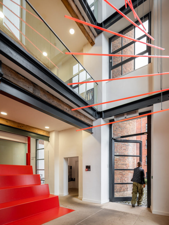 Suspended spiral light installation composed of intersecting illuminated linear elements arranged around a vertical stairwell void, with coloured light bars radiating and twisting through space to echo the geometry and movement of the surrounding architecture, creating layered reflections and depth within the entrance atrium.