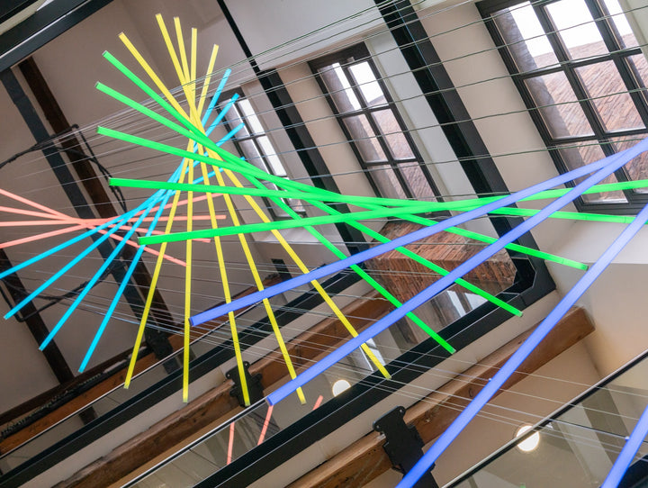 Suspended spiral light installation composed of intersecting illuminated linear elements arranged around a vertical stairwell void, with coloured light bars radiating and twisting through space to echo the geometry and movement of the surrounding architecture, creating layered reflections and depth within the entrance atrium.