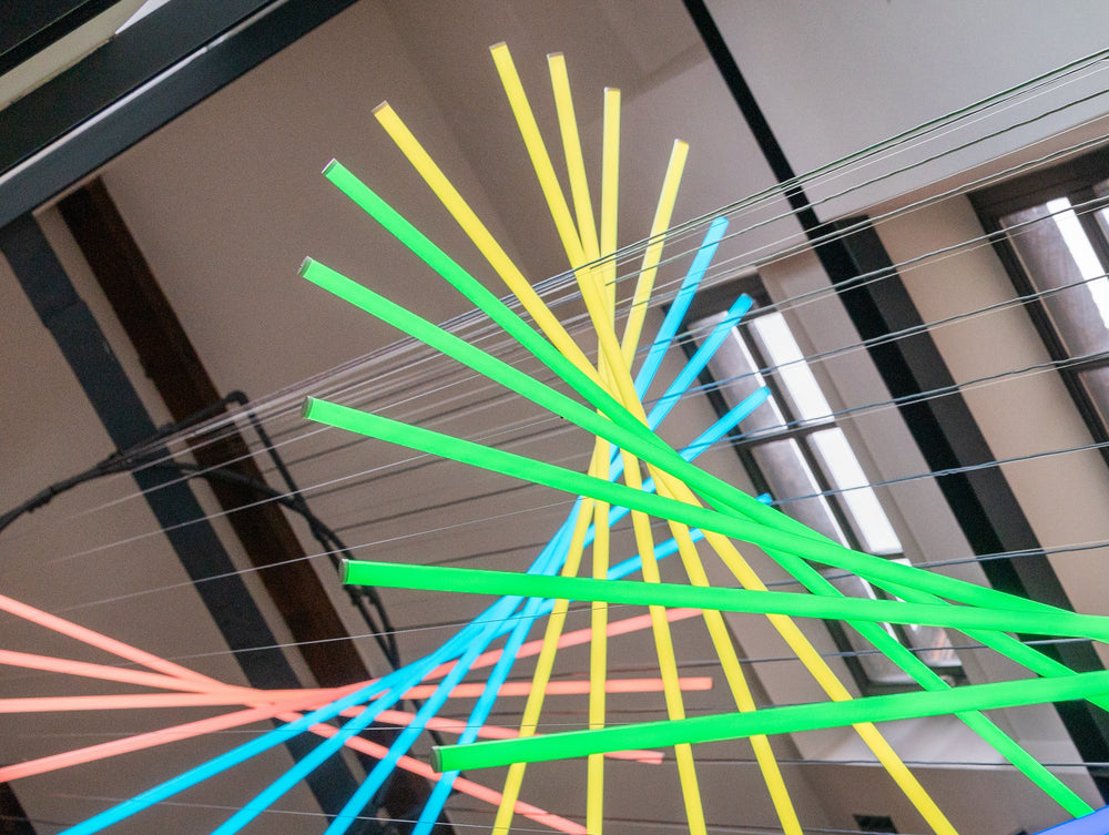 Suspended spiral light installation composed of intersecting illuminated linear elements arranged around a vertical stairwell void, with coloured light bars radiating and twisting through space to echo the geometry and movement of the surrounding architecture, creating layered reflections and depth within the entrance atrium.