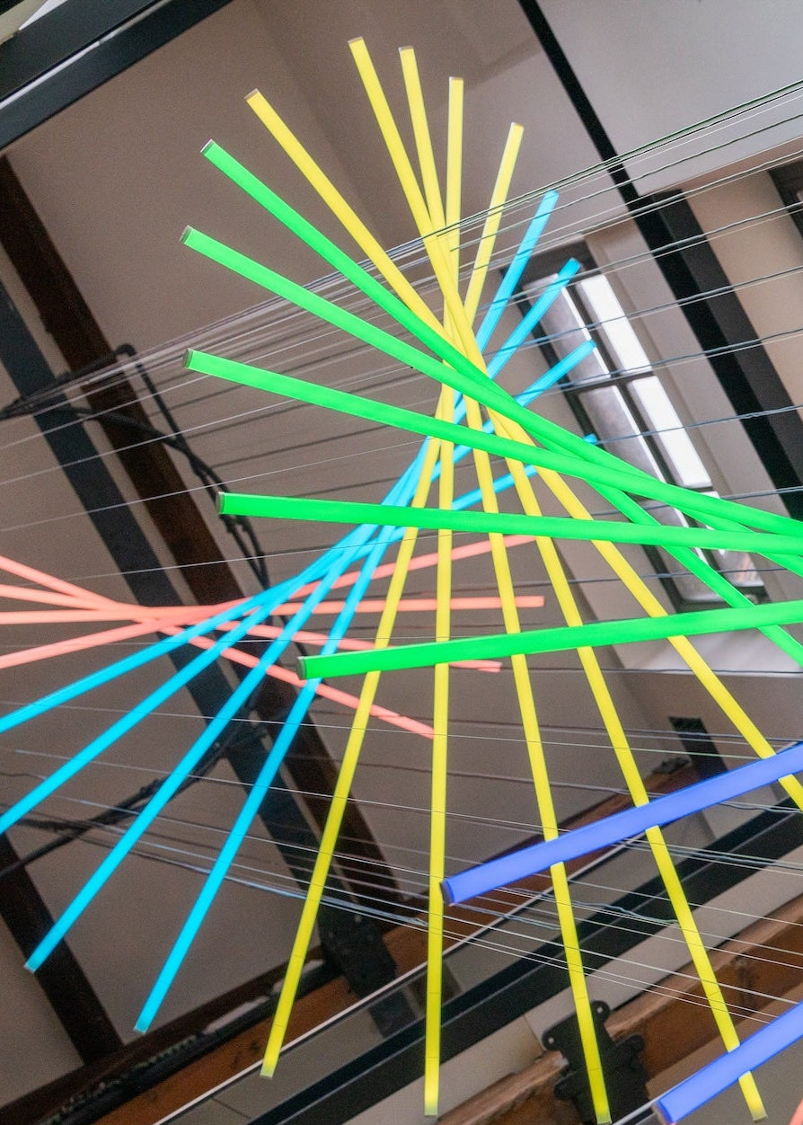 Suspended spiral light installation composed of intersecting illuminated linear elements arranged around a vertical stairwell void, with coloured light bars radiating and twisting through space to echo the geometry and movement of the surrounding architecture, creating layered reflections and depth within the entrance atrium.