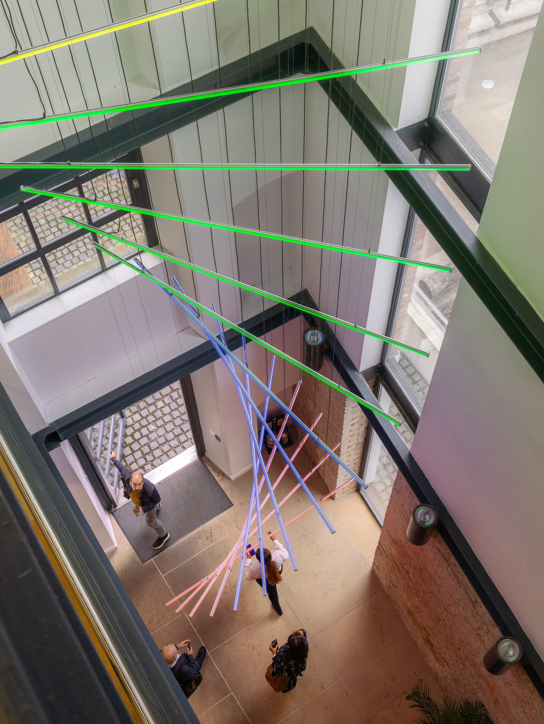 Suspended spiral light installation composed of intersecting illuminated linear elements arranged around a vertical stairwell void, with coloured light bars radiating and twisting through space to echo the geometry and movement of the surrounding architecture, creating layered reflections and depth within the entrance atrium.