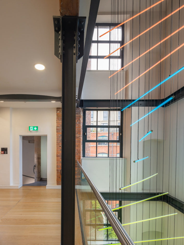 Suspended spiral light installation composed of intersecting illuminated linear elements arranged around a vertical stairwell void, with coloured light bars radiating and twisting through space to echo the geometry and movement of the surrounding architecture, creating layered reflections and depth within the entrance atrium.