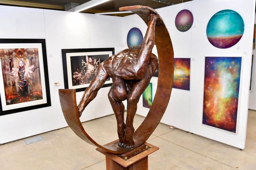 Weight of the World by Teresa Wells – a large, hand-cast bronze sculpture mounted on a tall steel base, depicting a nude male figure in a forward bend, gripping the ends of a sweeping curved bronze arc. The figure’s muscular back, bent legs, and tense feet convey physical strain and emotional weight, symbolising the burden of life’s pressures. Rich brown patina with subtle tonal variations enhances the anatomy and texture. Edition of 9, created 2019, 100cm × 45cm × 100cm, 100kg.