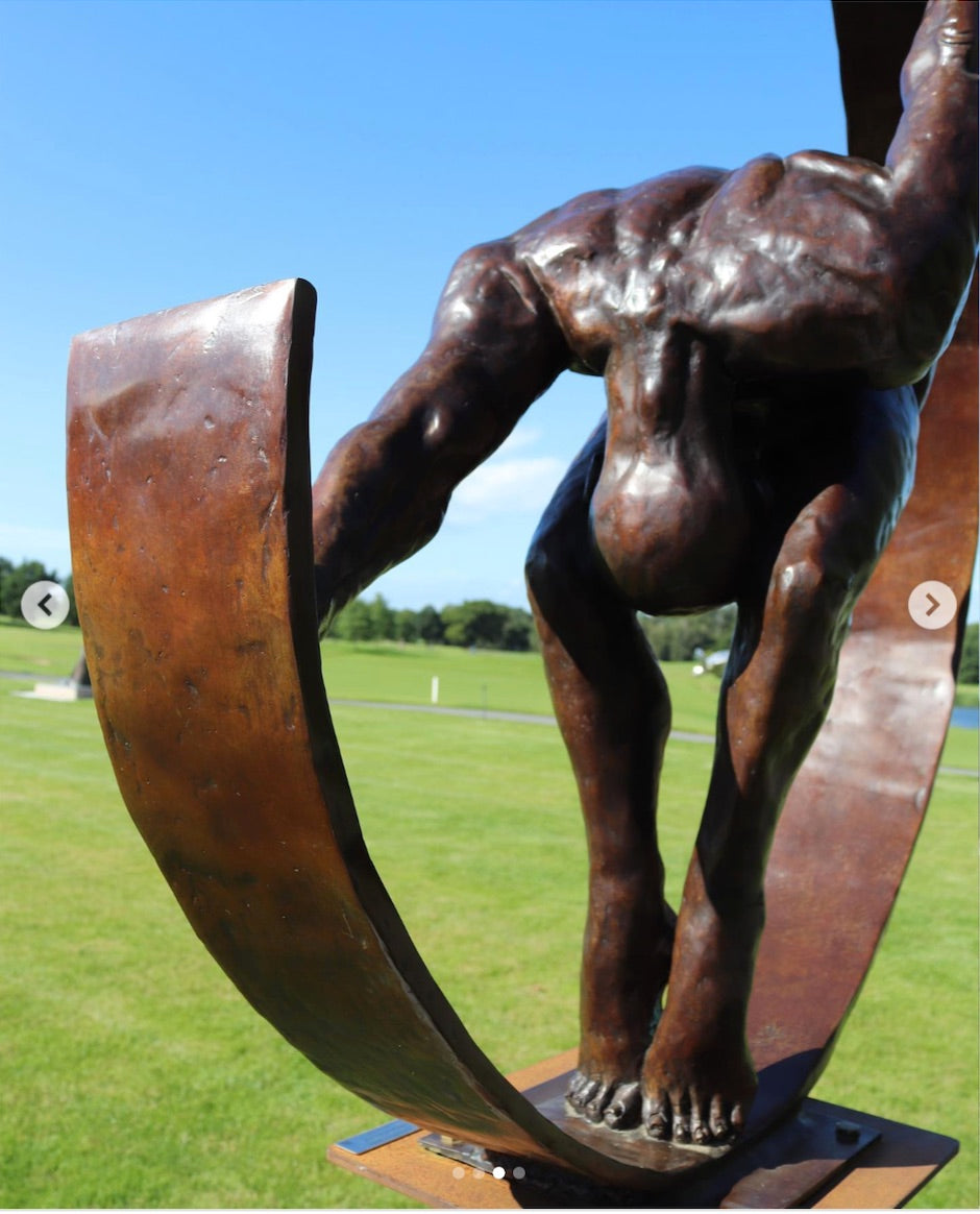 Weight of the World by Teresa Wells – a large, hand-cast bronze sculpture mounted on a tall steel base, depicting a nude male figure in a forward bend, gripping the ends of a sweeping curved bronze arc. The figure’s muscular back, bent legs, and tense feet convey physical strain and emotional weight, symbolising the burden of life’s pressures. Rich brown patina with subtle tonal variations enhances the anatomy and texture. Edition of 9, created 2019, 100cm × 45cm × 100cm, 100kg.