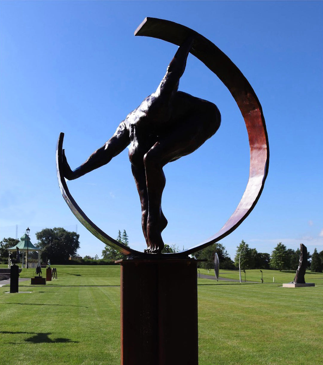 Weight of the World by Teresa Wells – a large, hand-cast bronze sculpture mounted on a tall steel base, depicting a nude male figure in a forward bend, gripping the ends of a sweeping curved bronze arc. The figure’s muscular back, bent legs, and tense feet convey physical strain and emotional weight, symbolising the burden of life’s pressures. Rich brown patina with subtle tonal variations enhances the anatomy and texture. Edition of 9, created 2019, 100cm × 45cm × 100cm, 100kg.